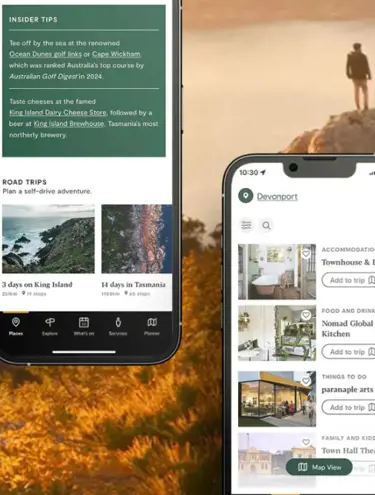 Two phone screens show the Discover Tasmania app display. The upper-left screen shows a series of insider tips and road trips, with photos of Tasmanian destinations and itinerary headlines. The lower-right phone displays Devonport accommodation and food and drink listings, next to photos of these businesses and a button asking to "Add to trip". Behind these two phones as the background image is a man standing on a rock in the golden sunlight, high above the water.