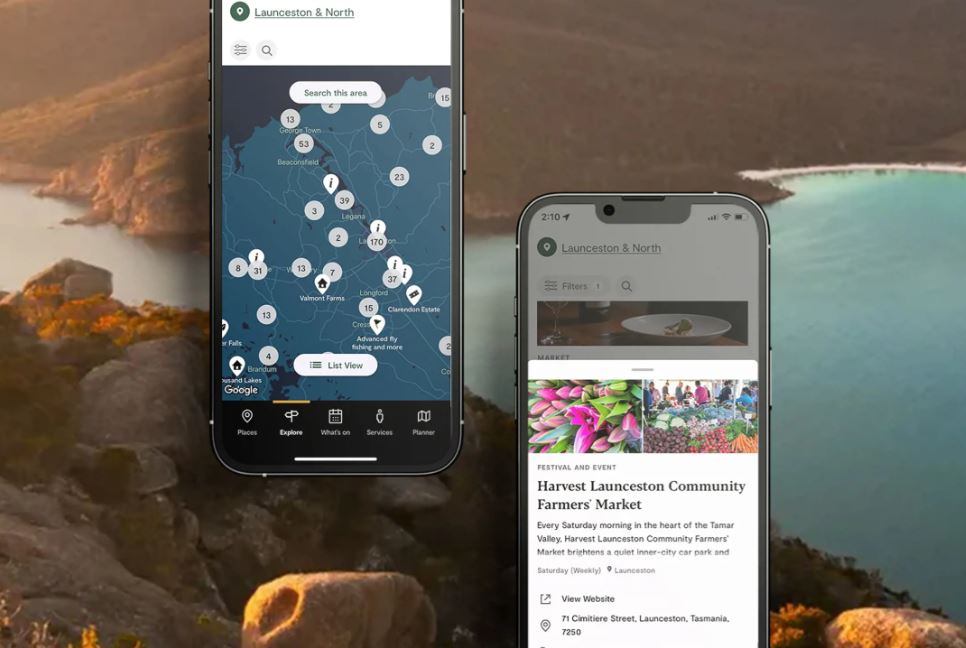 A scenic backdrop shows turquoise sea surrounded by rocky headlands and tree-lined hillsides. In the foreground are two phones displaying the Discover Tasmania ap. The upper-left phone displays a map of "Launceston & north" with numbered stops along the way. The lower-right phone displays photos of the Harvest Launceston Community Farmers' Market, with a short description of the event, its address, its location, information stating it is open "Saturday (Weekly)" and a link to view the business' website.