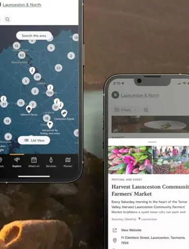 A scenic backdrop shows turquoise sea surrounded by rocky headlands and tree-lined hillsides. In the foreground are two phones displaying the Discover Tasmania ap. The upper-left phone displays a map of "Launceston & north" with numbered stops along the way. The lower-right phone displays photos of the Harvest Launceston Community Farmers' Market, with a short description of the event, its address, its location, information stating it is open "Saturday (Weekly)" and a link to view the business' website.