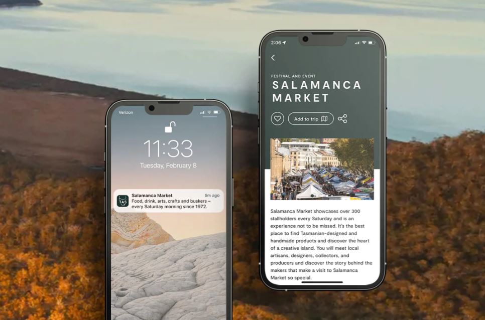 Two phone screens are displayed in front of a background image of golden bushland and hillsides with a grey sky. The phone screen on the left displays a Discover Tasmania app notification that reads: "Salamanca Market: Food, drink, arts, crafts and buskers - every Saturday morning since 1972." The right-hand phone screen displays the Salamanca Market business listing on the Discover Tasmania app, showing a photo of the market followed by a description of the event. At the top underneath the "Salamanca Market" heading, there's a love-heart button, an "Add to trip" button and a link-share button.