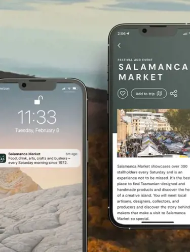 Two phone screens are displayed in front of a background image of golden bushland and hillsides with a grey sky. The phone screen on the left displays a Discover Tasmania app notification that reads: "Salamanca Market: Food, drink, arts, crafts and buskers - every Saturday morning since 1972." The right-hand phone screen displays the Salamanca Market business listing on the Discover Tasmania app, showing a photo of the market followed by a description of the event. At the top underneath the "Salamanca Market" heading, there's a love-heart button, an "Add to trip" button and a link-share button.