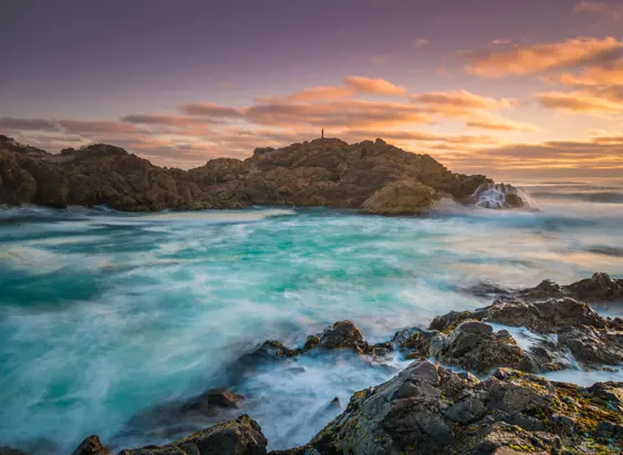 A person silhouetted on a rocky headland with waves breaking into a turquoise bay at Trial Harbour, Tasmania's West Coast