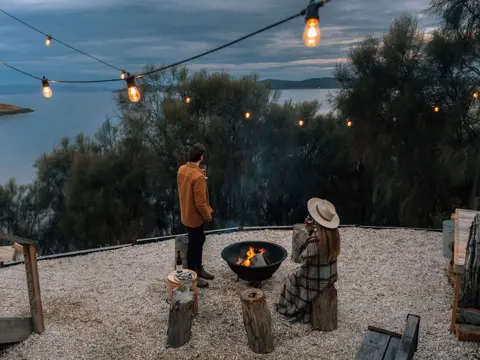 Guests by a firepit and string lights with ocean views at Driftwood Cabin, Primrose Sands, Tasmania