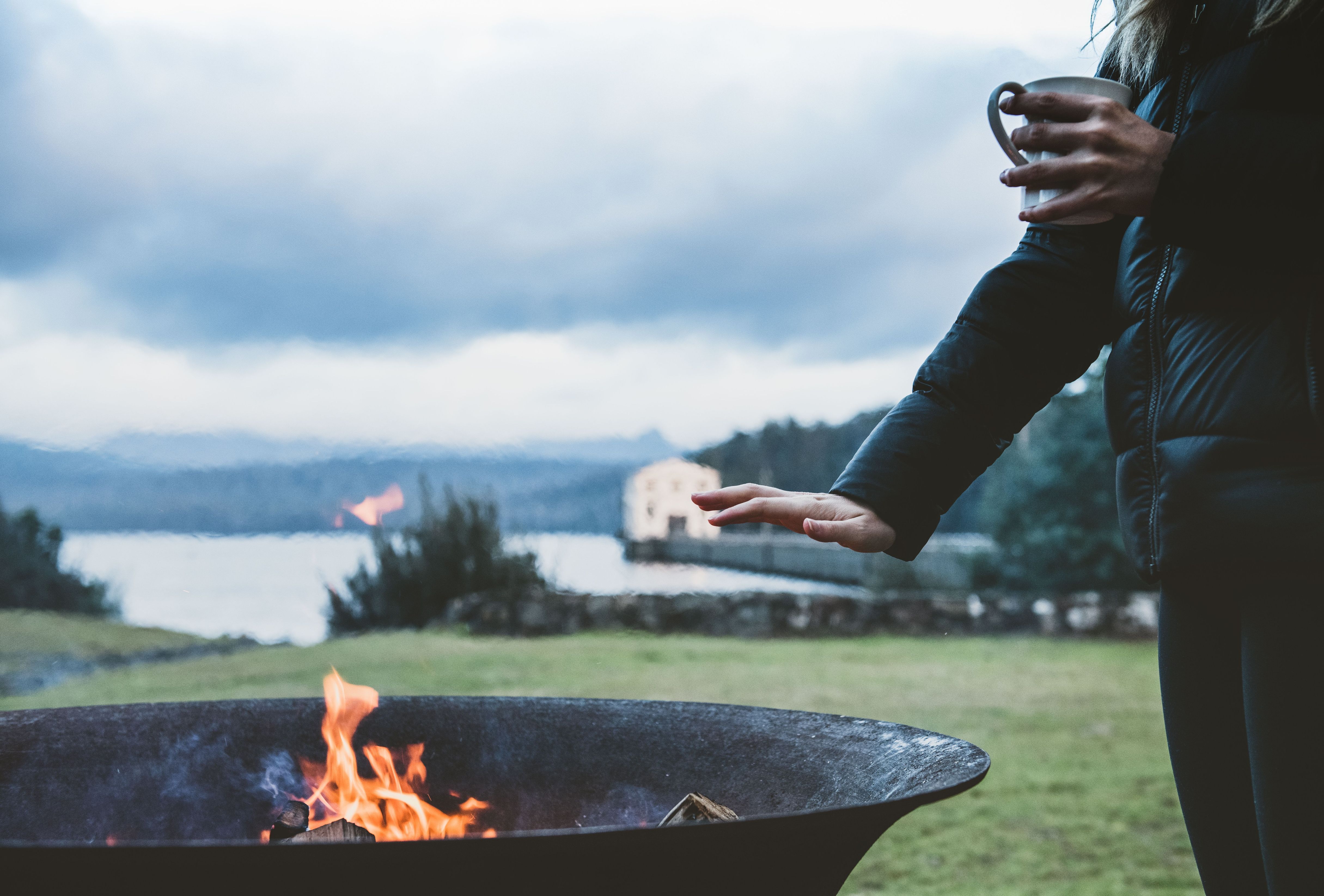 A guest with a mug warming hands by a firepit, Pumphouse Point visible in the background, Lake St Clair, Tasmania