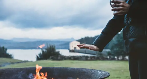 A guest with a mug warming hands by a firepit, Pumphouse Point visible in the background, Lake St Clair, Tasmania