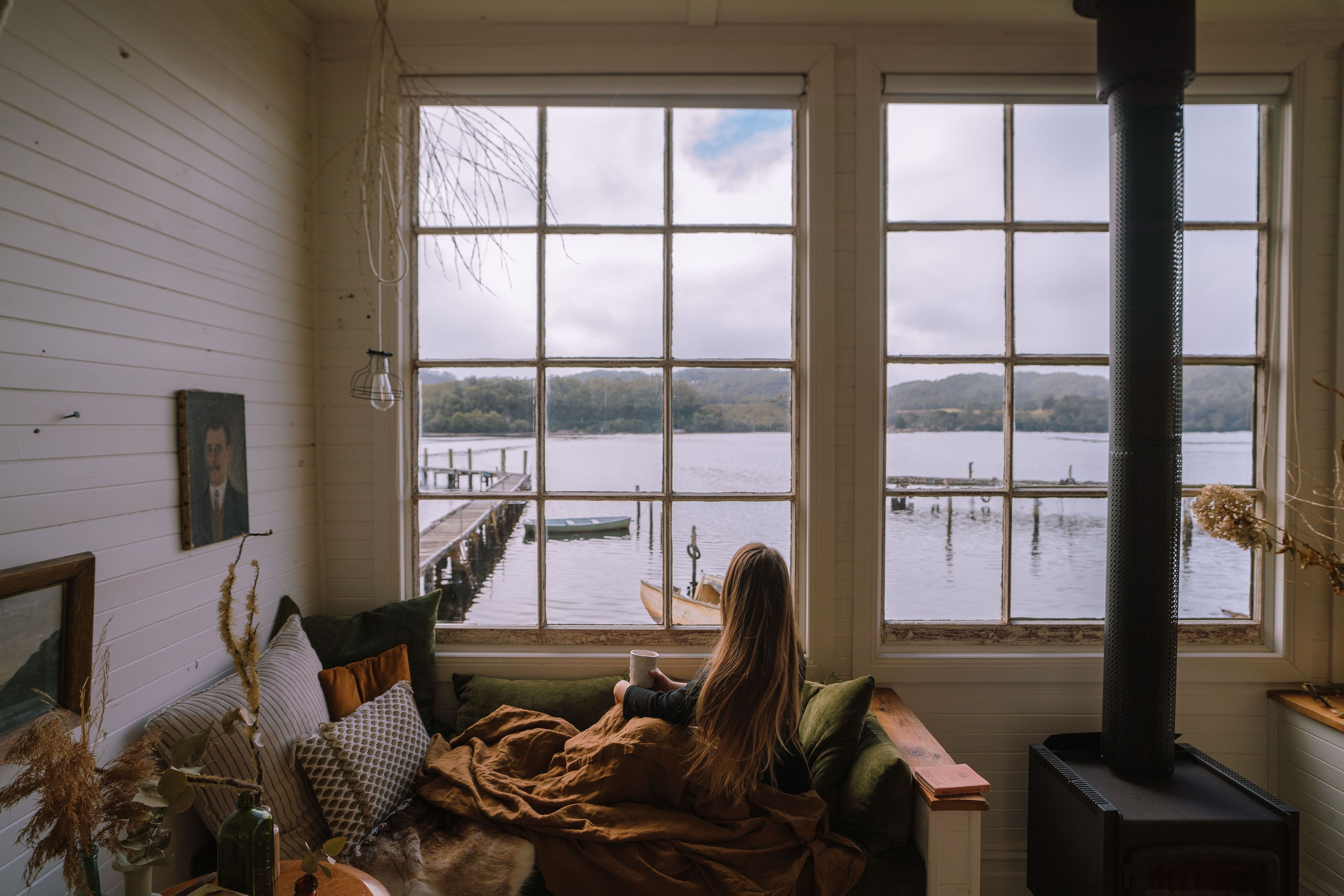 A guest with a mug looking through windows toward the water and jetty at Captain's Rest, Strahan on Tasmania's West Coast