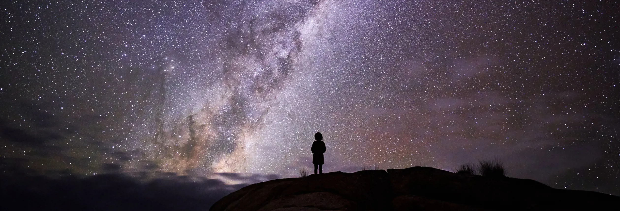 Person silhouetted on a rocky outcrop beneath the Milky Way at larapuna / Bay of Fires area, Tasmania's East Coast