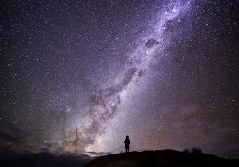Person silhouetted on a rocky outcrop beneath the Milky Way at larapuna / Bay of Fires area, Tasmania's East Coast