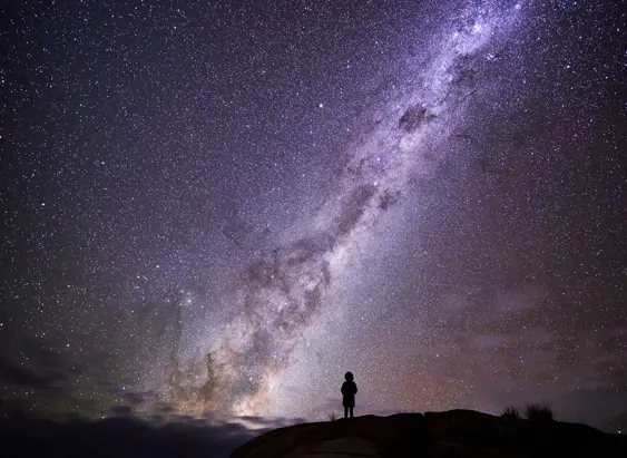 Person silhouetted on a rocky outcrop beneath the Milky Way at larapuna / Bay of Fires area, Tasmania's East Coast