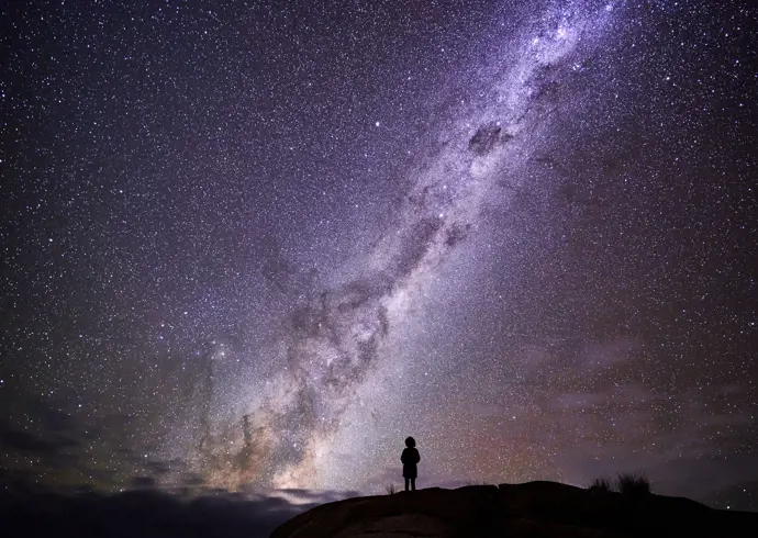 Person silhouetted on a rocky outcrop beneath the Milky Way at larapuna / Bay of Fires area, Tasmania's East Coast
