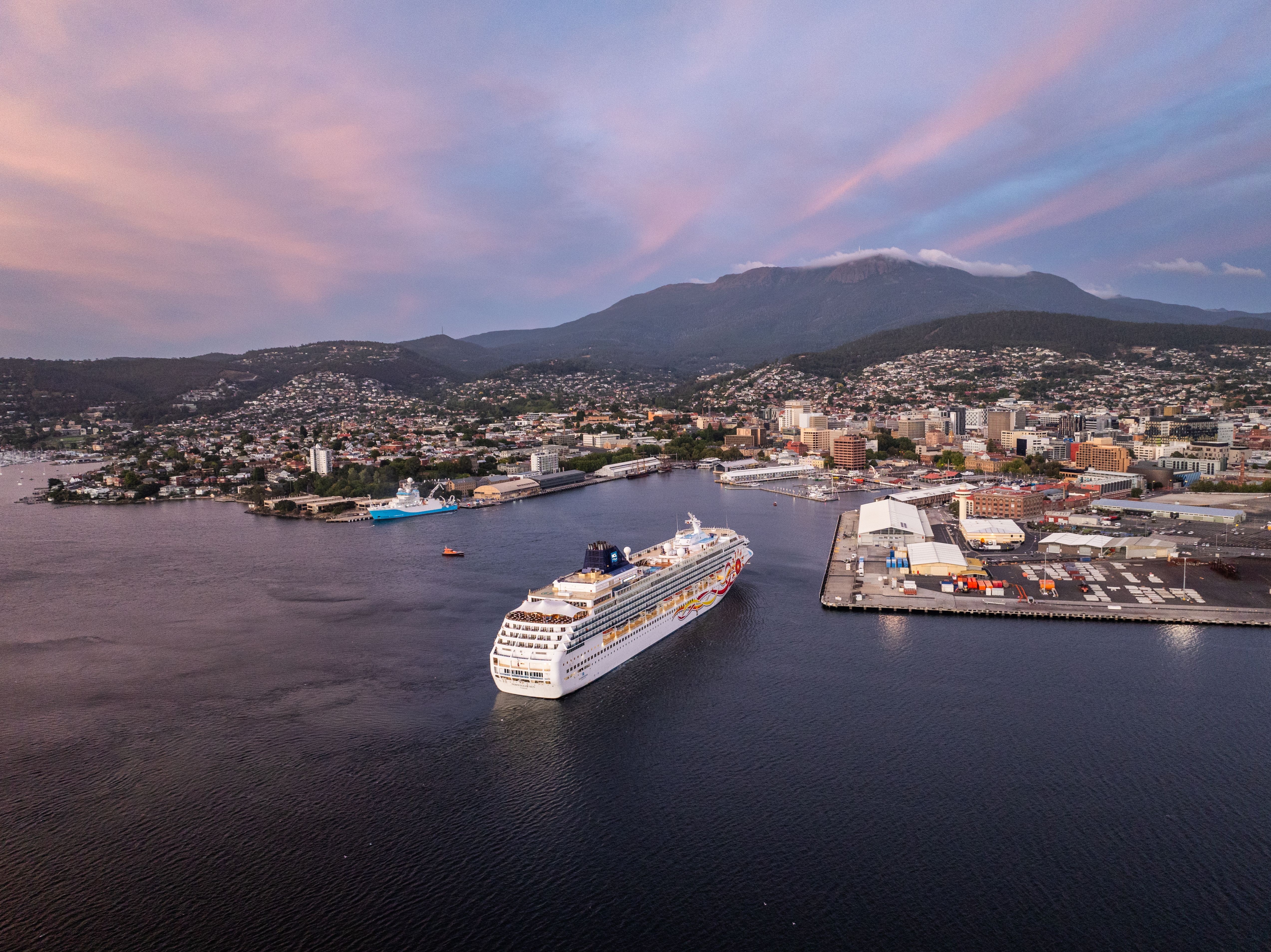 A large cruise ship gliding into a harbour at dusk, with a hillside town and soft pink sunset sky forming the backdrop.