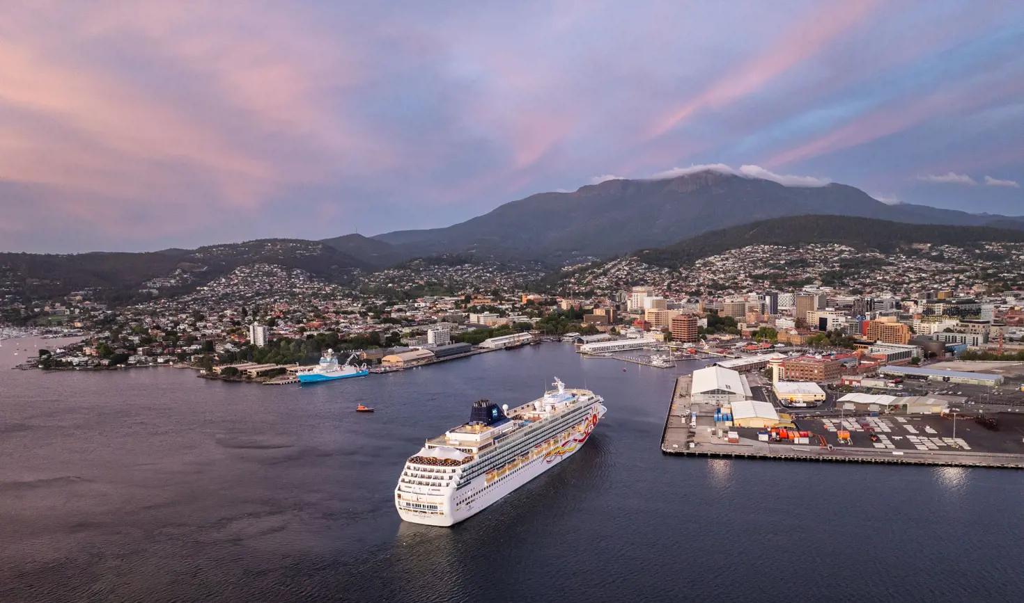 A large cruise ship gliding into a harbour at dusk, with a hillside town and soft pink sunset sky forming the backdrop.