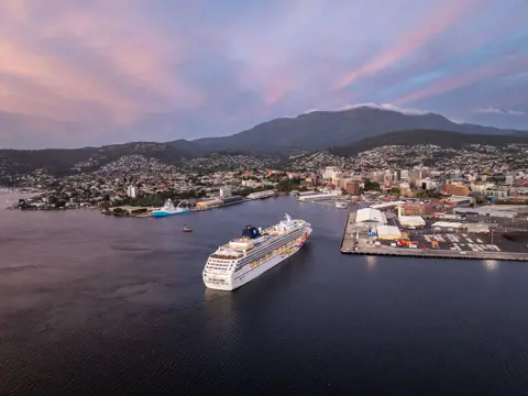 A large cruise ship gliding into a harbour at dusk, with a hillside town and soft pink sunset sky forming the backdrop.