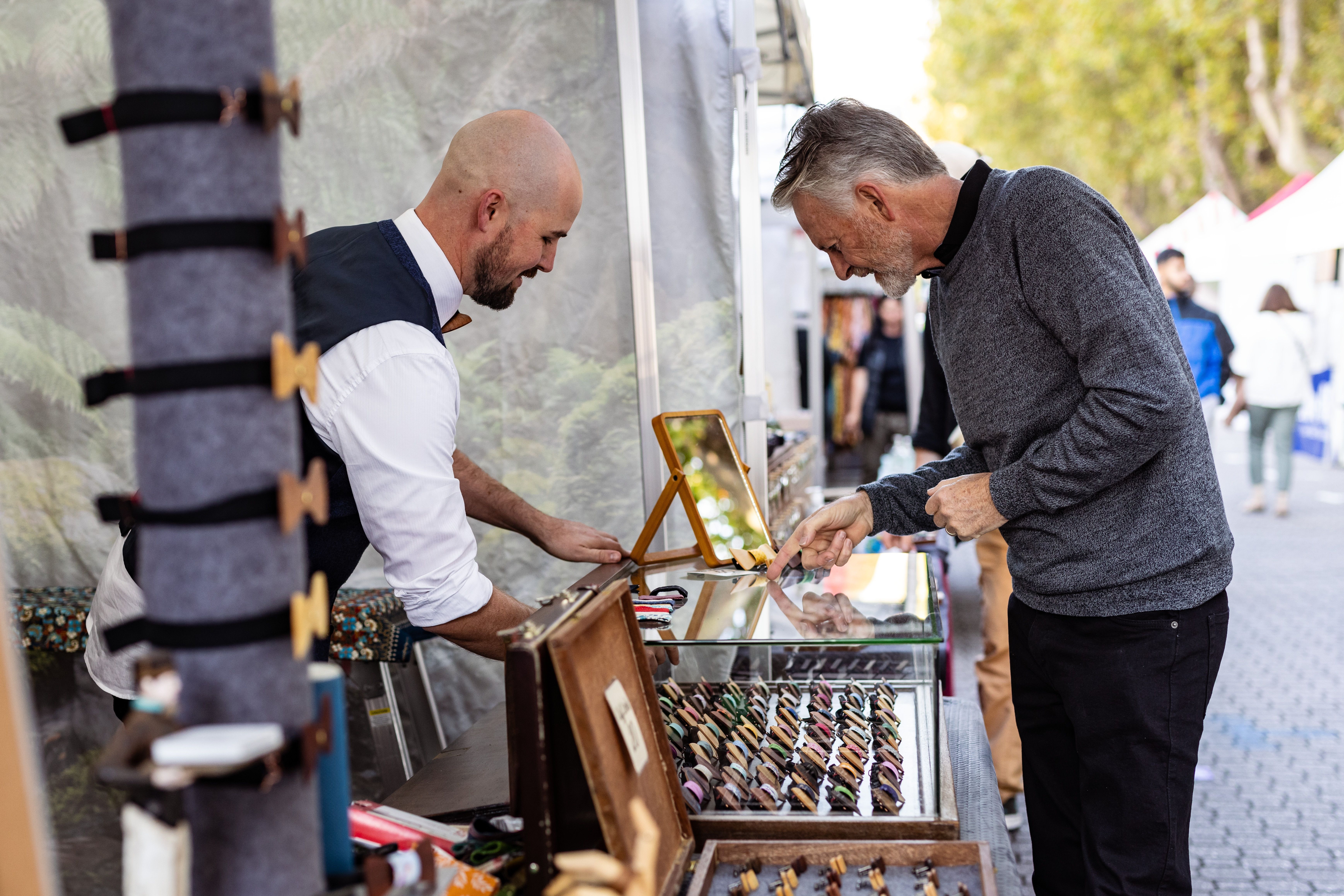 A stallholder in a white shirt and waistcoat serving a customer across a market stall displaying a selection of small products in rows, with a bustling outdoor market visible in the background.