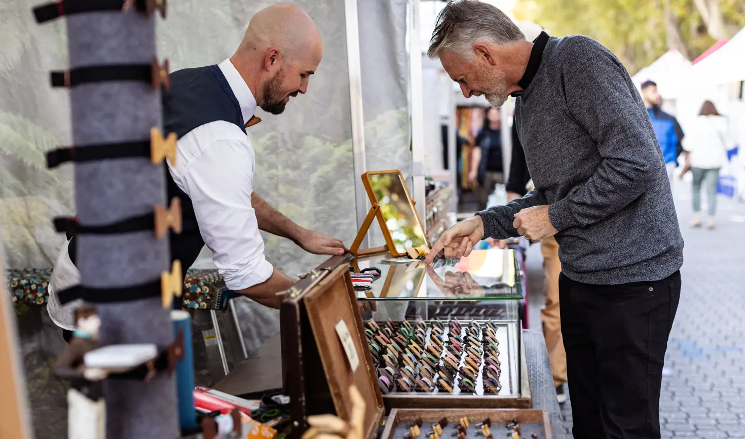 A stallholder in a white shirt and waistcoat serving a customer across a market stall displaying a selection of small products in rows, with a bustling outdoor market visible in the background.