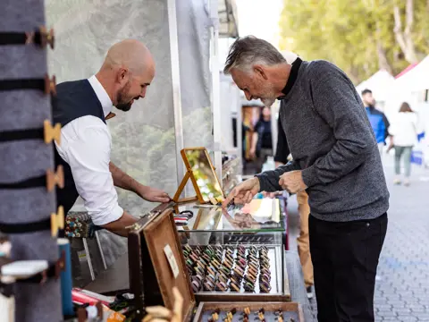 A stallholder in a white shirt and waistcoat serving a customer across a market stall displaying a selection of small products in rows, with a bustling outdoor market visible in the background.
