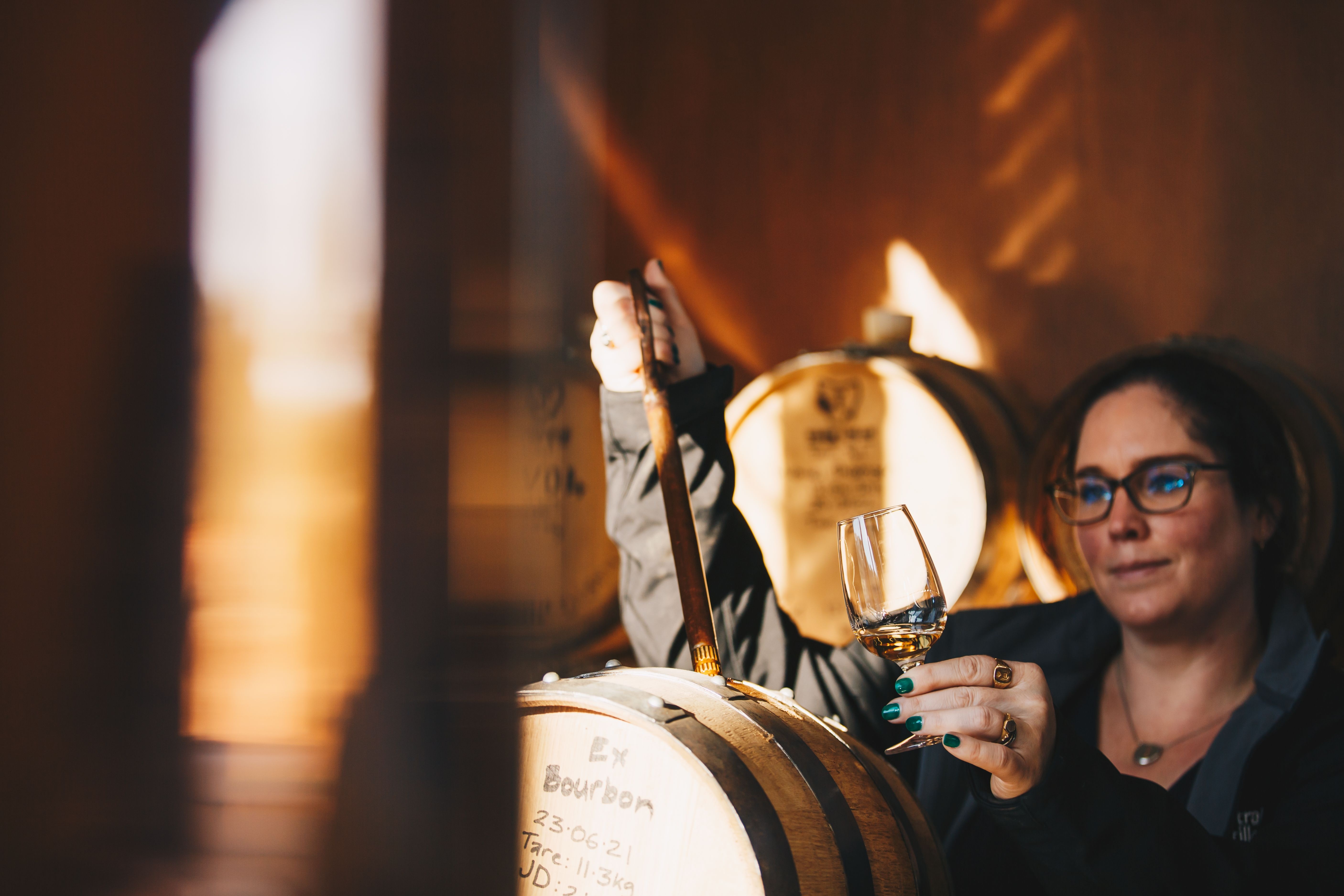 A woman holding a glass of amber spirit up to the light while drawing a sample from an oak barrel in a warmly lit distillery, rows of barrels visible in the soft golden background.