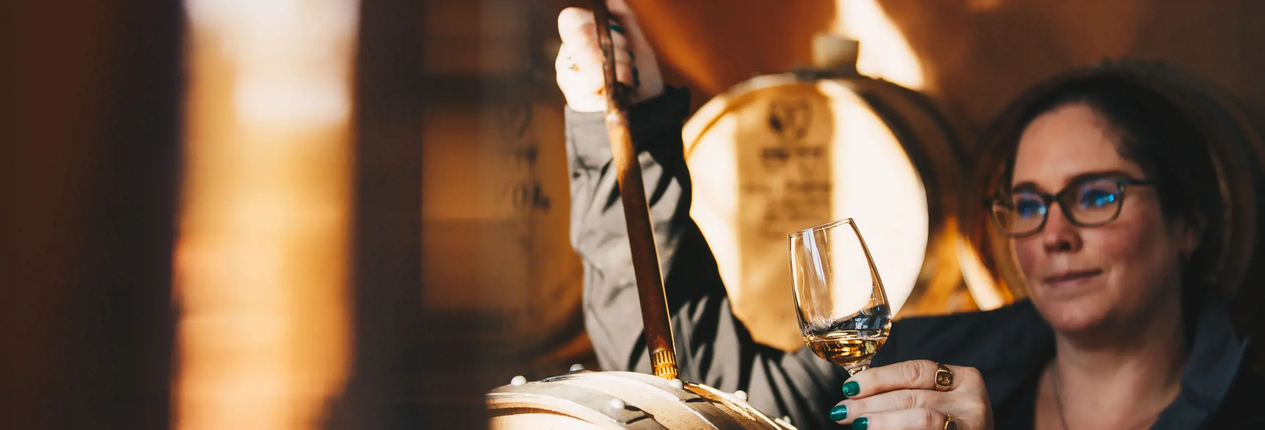 A woman holding a glass of amber spirit up to the light while drawing a sample from an oak barrel in a warmly lit distillery, rows of barrels visible in the soft golden background.