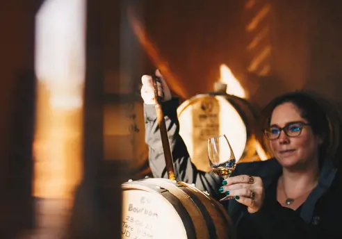 A woman holding a glass of amber spirit up to the light while drawing a sample from an oak barrel in a warmly lit distillery, rows of barrels visible in the soft golden background.