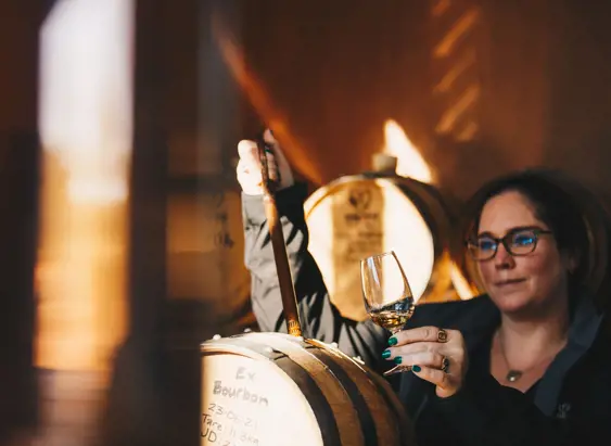 A woman holding a glass of amber spirit up to the light while drawing a sample from an oak barrel in a warmly lit distillery, rows of barrels visible in the soft golden background.
