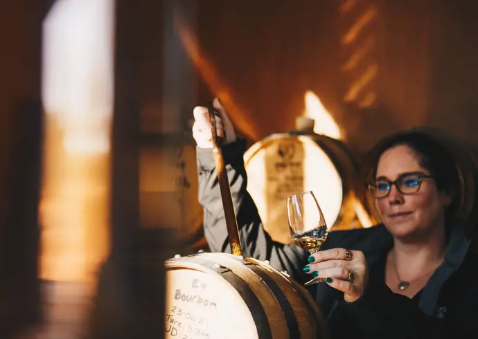 A woman holding a glass of amber spirit up to the light while drawing a sample from an oak barrel in a warmly lit distillery, rows of barrels visible in the soft golden background.