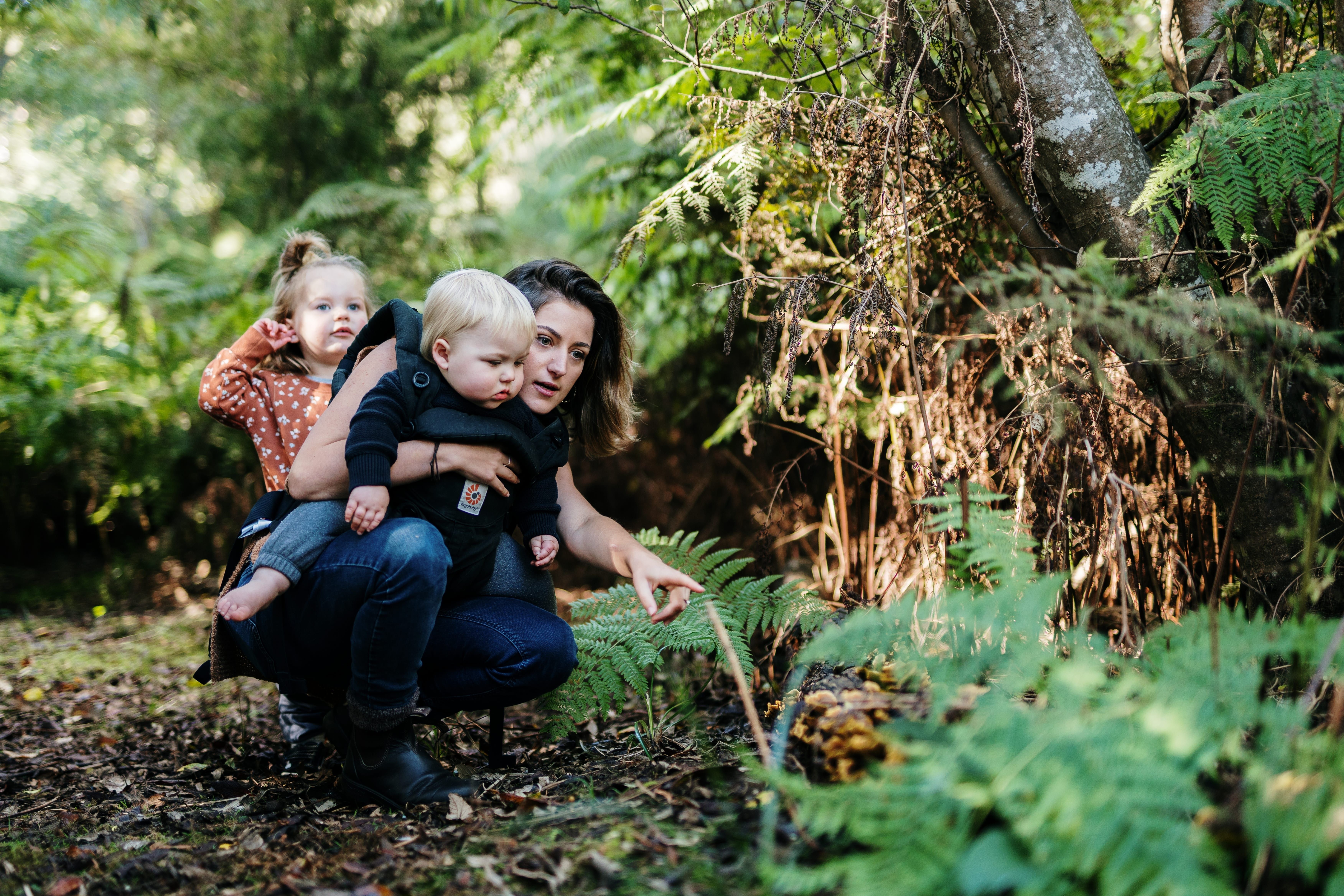 A woman crouching beside a fern in a mossy forest, holding a toddler on her hip and pointing something out to a young child standing nearby, all three engaged in quiet discovery among the lush green undergrowth.