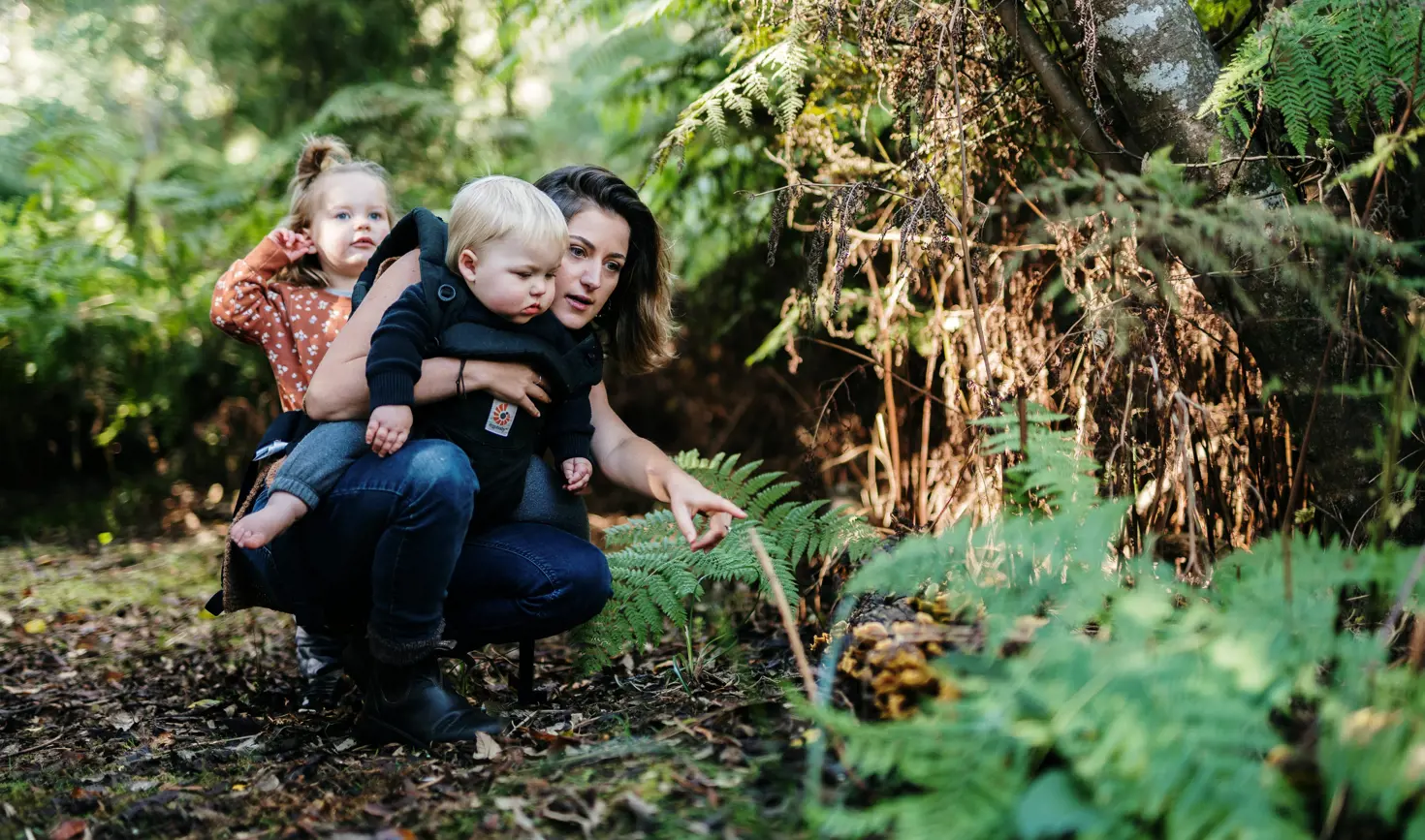 A woman crouching beside a fern in a mossy forest, holding a toddler on her hip and pointing something out to a young child standing nearby, all three engaged in quiet discovery among the lush green undergrowth.