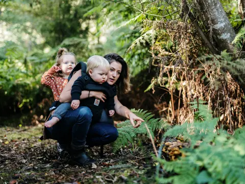 A woman crouching beside a fern in a mossy forest, holding a toddler on her hip and pointing something out to a young child standing nearby, all three engaged in quiet discovery among the lush green undergrowth.