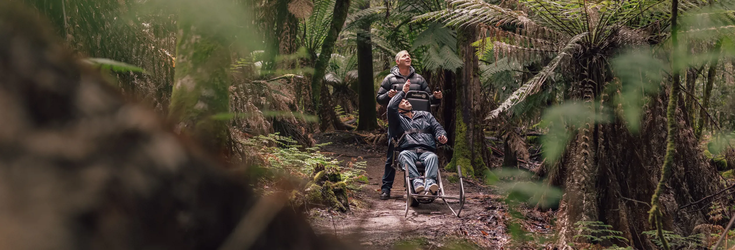 A person being carefully assisted along a bush trail in an all-terrain wheelchair, moving through the forest with support from a companion.