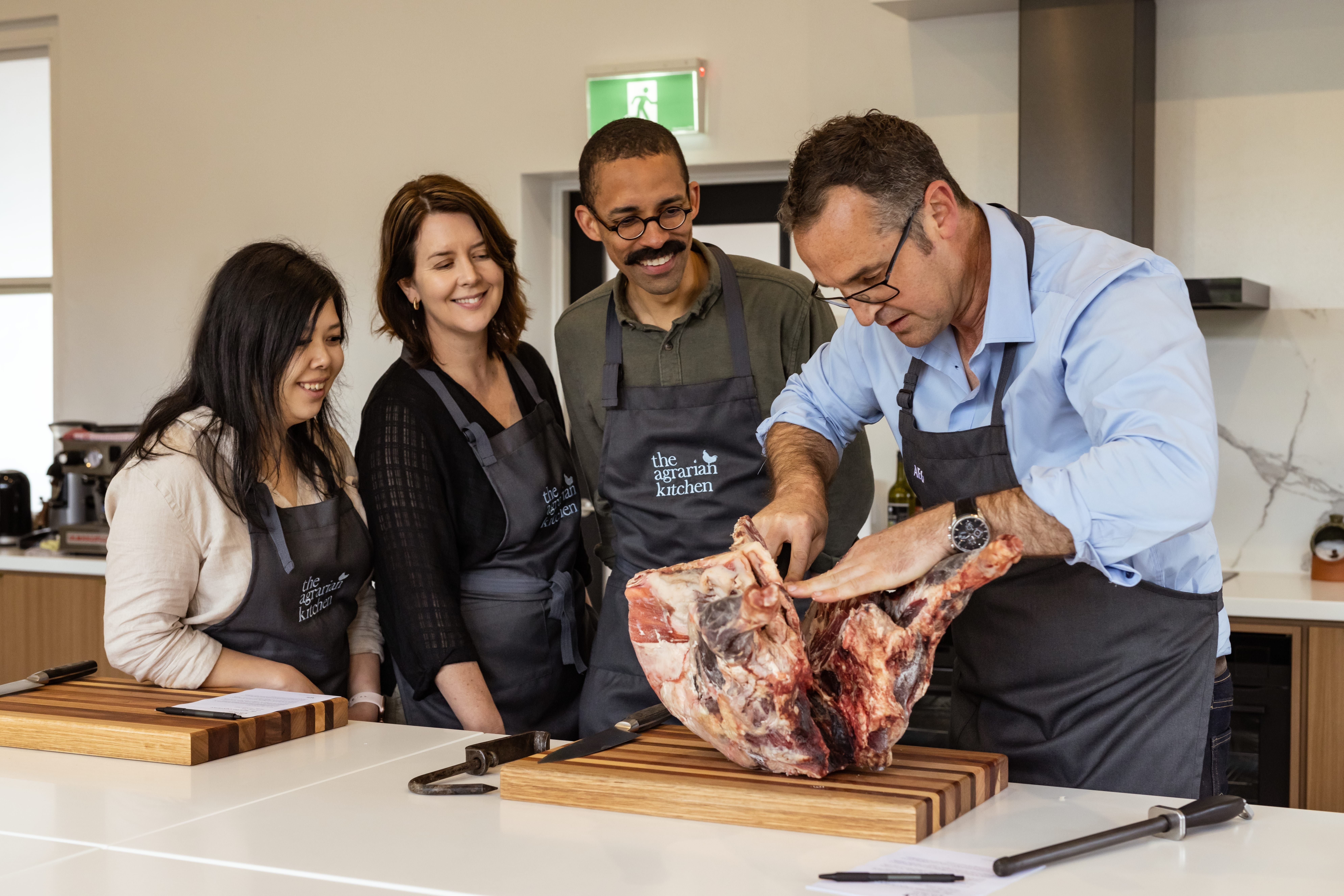 A chef in a blue shirt demonstrating how to break down a large cut of meat on a wooden board, with three attentive participants in aprons watching closely in a bright, modern kitchen.
