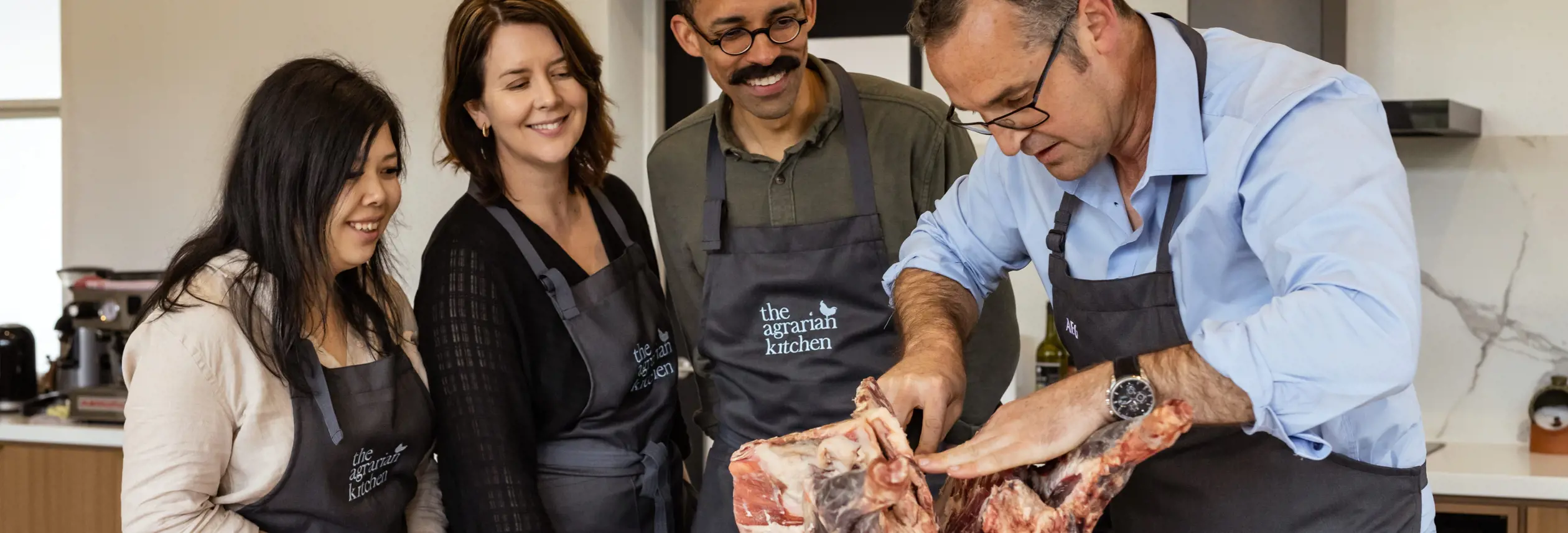 A chef in a blue shirt demonstrating how to break down a large cut of meat on a wooden board, with three attentive participants in aprons watching closely in a bright, modern kitchen.