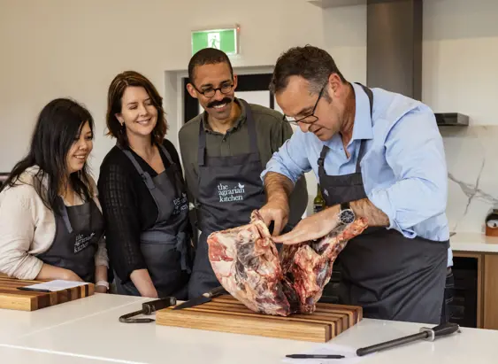 A chef in a blue shirt demonstrating how to break down a large cut of meat on a wooden board, with three attentive participants in aprons watching closely in a bright, modern kitchen.