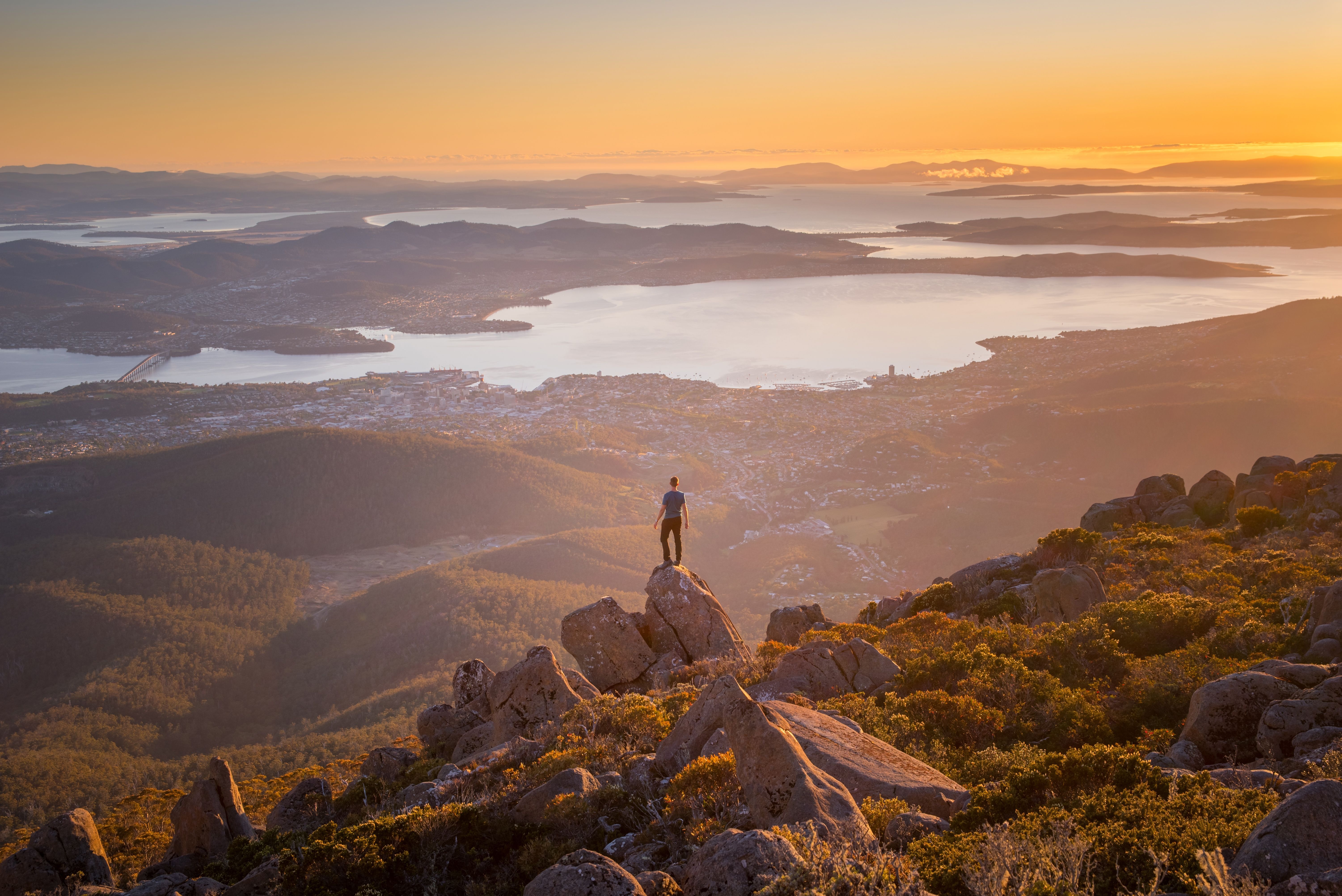 A lone figure standing on a rocky summit at sunset, looking out over a sweeping panorama of a city, winding river and distant bays bathed in warm golden light.