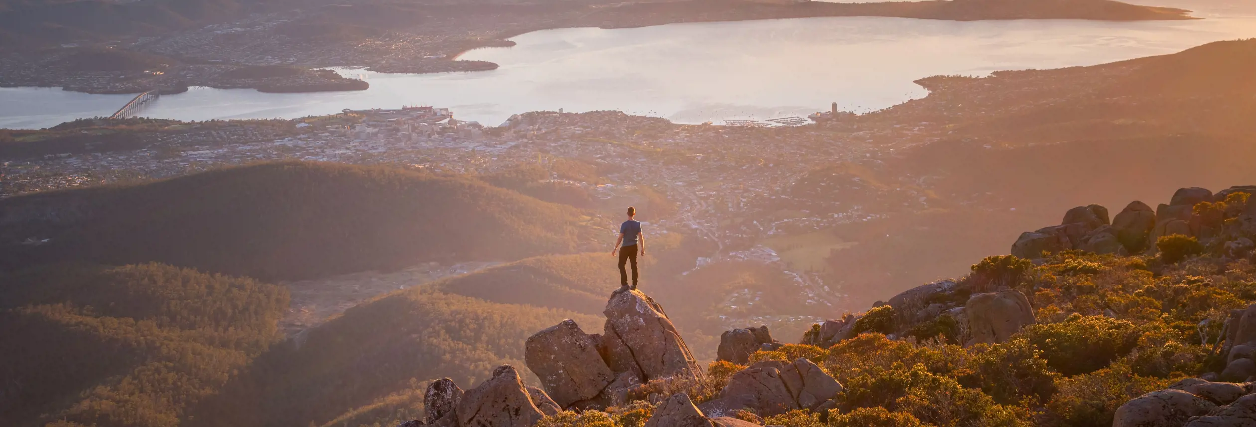 A lone figure standing on a rocky summit at sunset, looking out over a sweeping panorama of a city, winding river and distant bays bathed in warm golden light.