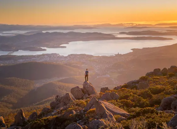 A lone figure standing on a rocky summit at sunset, looking out over a sweeping panorama of a city, winding river and distant bays bathed in warm golden light.