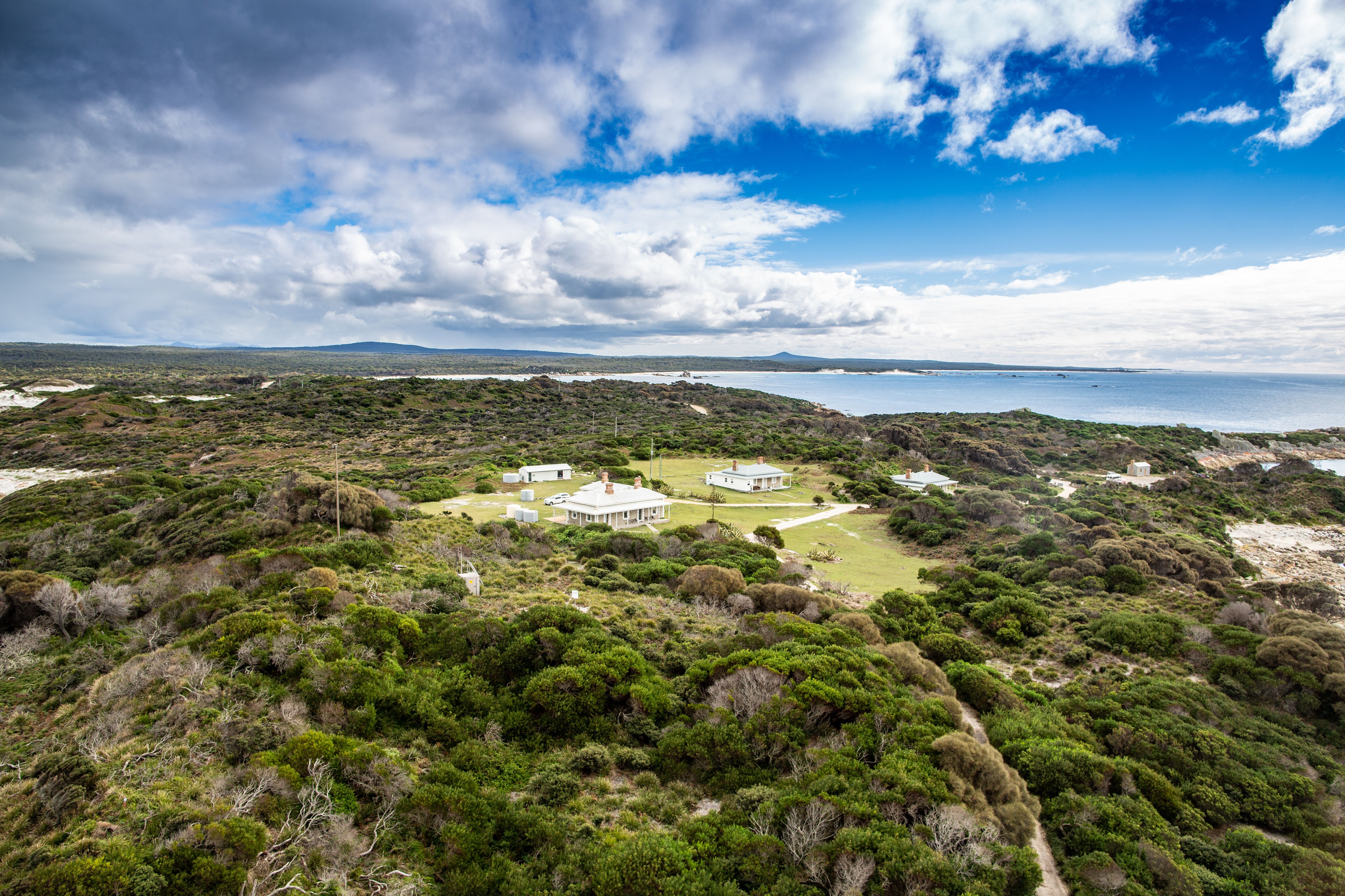 An aerial view of a cluster of white heritage buildings nestled in low coastal scrubland, with sandy beaches, a calm bay and distant headlands stretching out beneath a bright blue sky with scattered clouds.