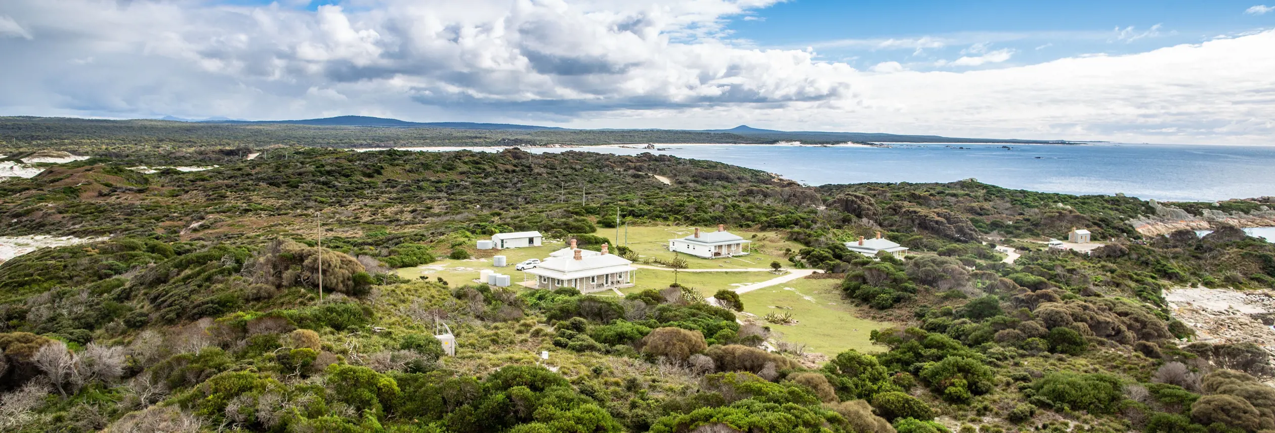 An aerial view of a cluster of white heritage buildings nestled in low coastal scrubland, with sandy beaches, a calm bay and distant headlands stretching out beneath a bright blue sky with scattered clouds.