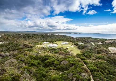 An aerial view of a cluster of white heritage buildings nestled in low coastal scrubland, with sandy beaches, a calm bay and distant headlands stretching out beneath a bright blue sky with scattered clouds.