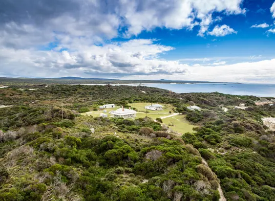 An aerial view of a cluster of white heritage buildings nestled in low coastal scrubland, with sandy beaches, a calm bay and distant headlands stretching out beneath a bright blue sky with scattered clouds.