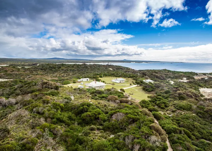An aerial view of a cluster of white heritage buildings nestled in low coastal scrubland, with sandy beaches, a calm bay and distant headlands stretching out beneath a bright blue sky with scattered clouds.