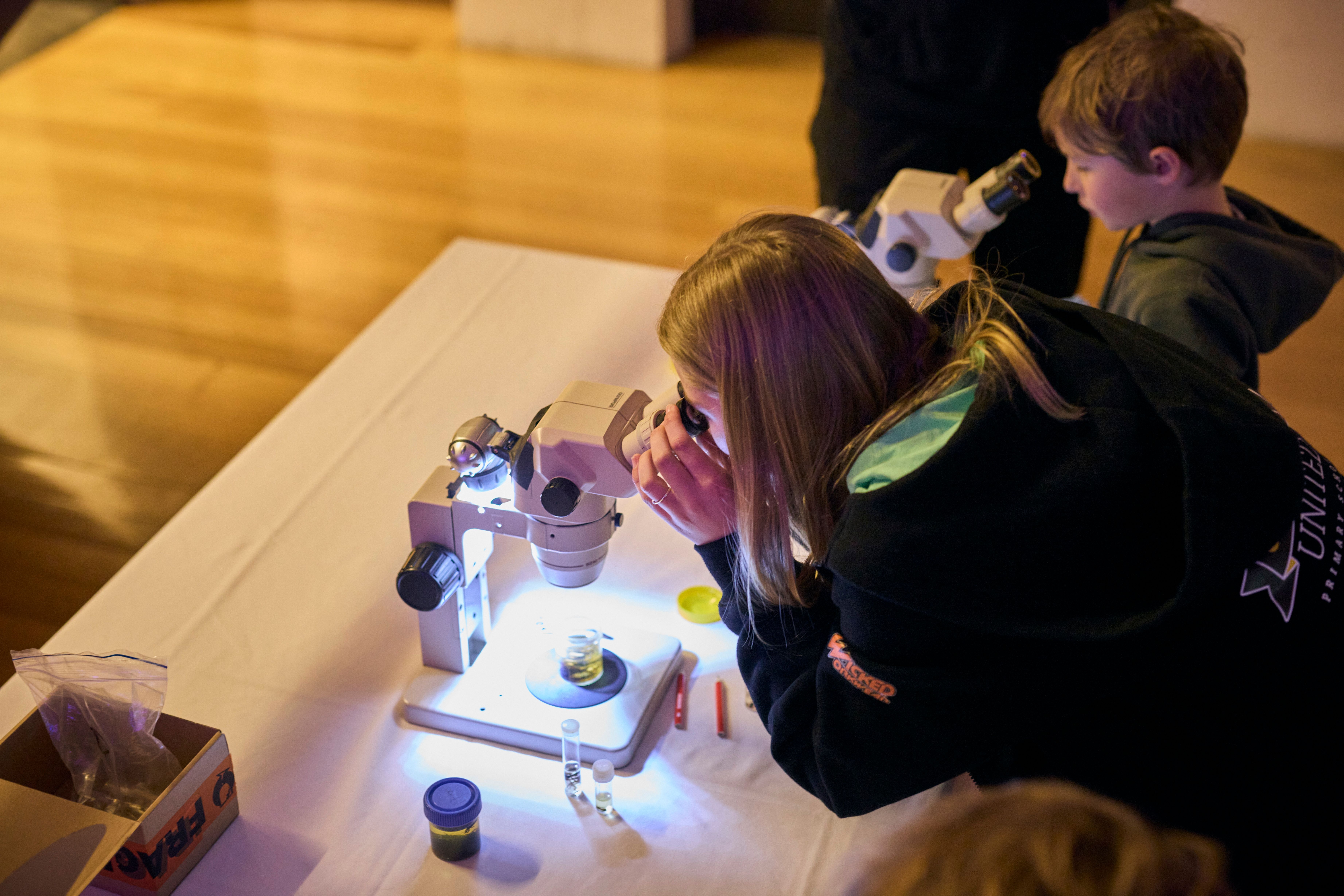 Two children bent over illuminated microscopes on a table, engrossed in examining specimens in what appears to be a hands-on science activity or educational workshop.