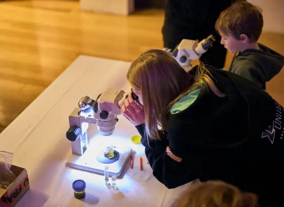 Two children bent over illuminated microscopes on a table, engrossed in examining specimens in what appears to be a hands-on science activity or educational workshop.