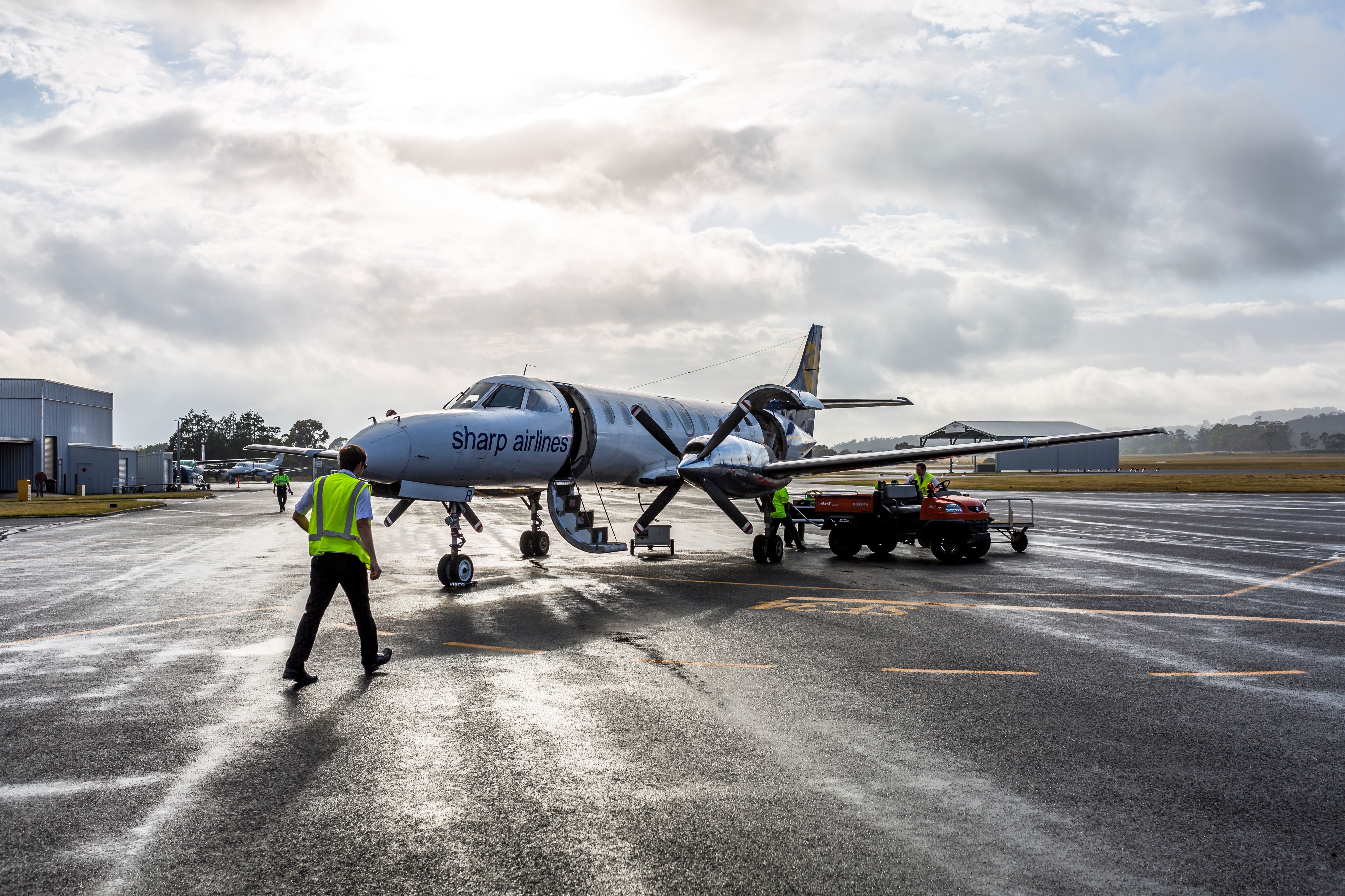 Ground crew in high-visibility vests working around a small regional turboprop aircraft on a wet tarmac, with airport buildings and a dramatic cloudy sky visible in the background.
