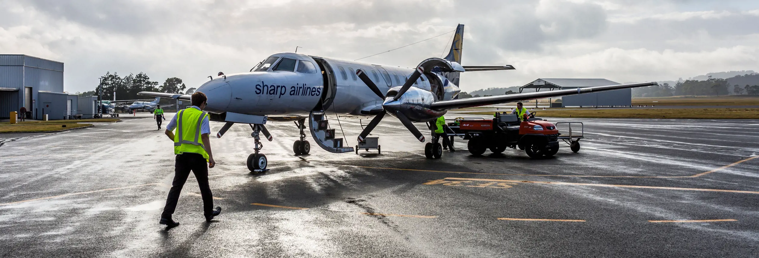 Ground crew in high-visibility vests working around a small regional turboprop aircraft on a wet tarmac, with airport buildings and a dramatic cloudy sky visible in the background.