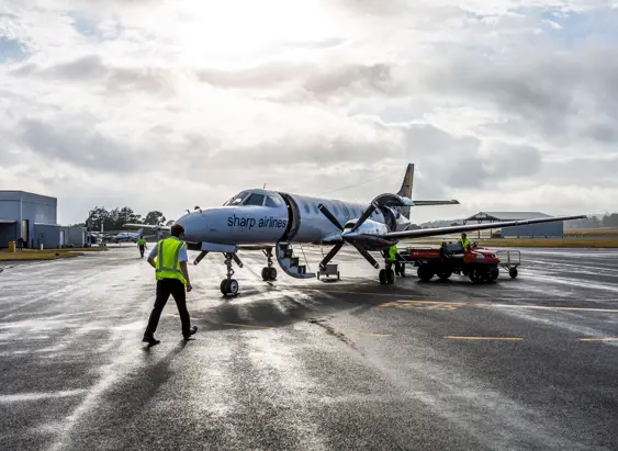 Ground crew in high-visibility vests working around a small regional turboprop aircraft on a wet tarmac, with airport buildings and a dramatic cloudy sky visible in the background.