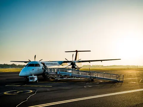 A small regional aircraft on the tarmac of an airport, with green rolling hills visible in the background beyond the runway.