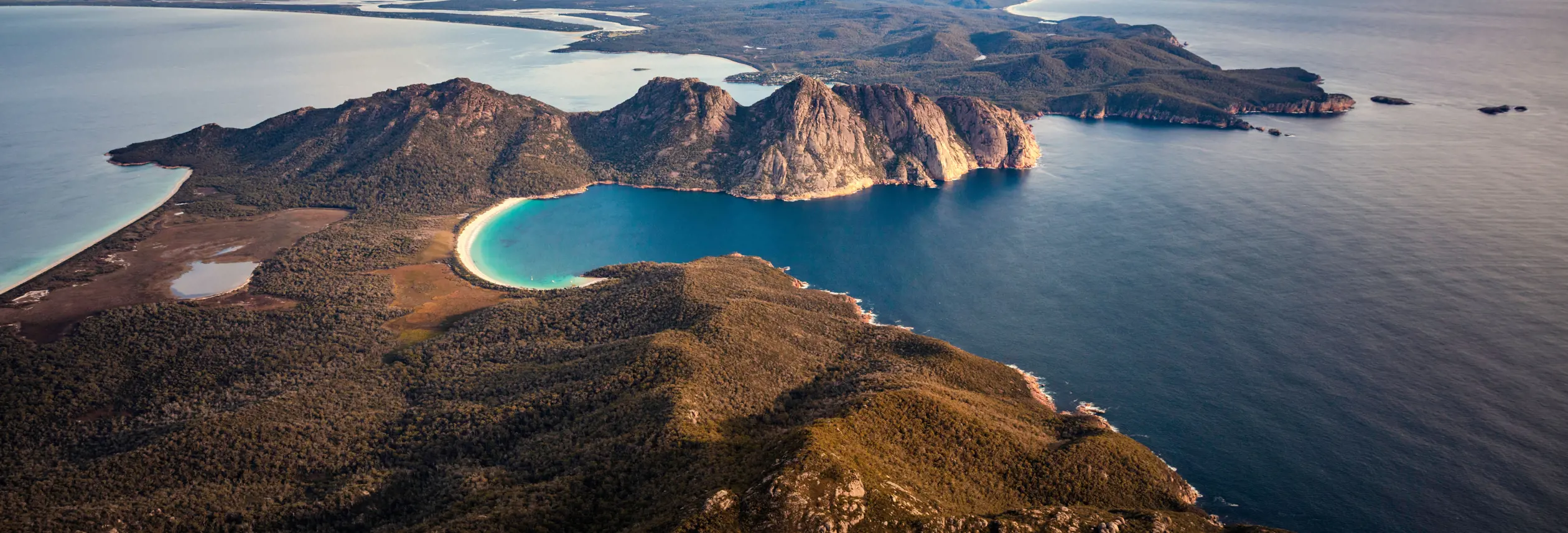 An aerial view of dramatic coastal cliffs dropping away to a crescent of white sand beach and vivid turquoise water below.