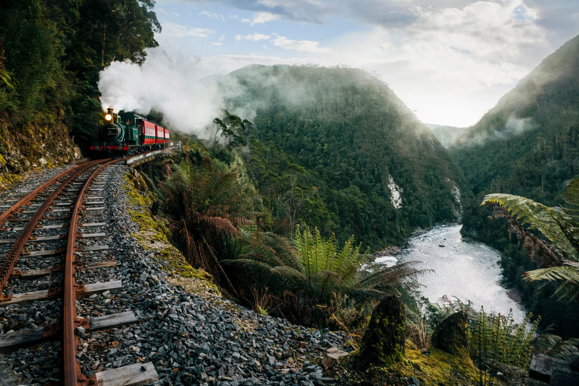 A vintage steam train rounding a curve on a narrow mountain railway, with white steam billowing into a lush gorge of ferns and moss-covered cliffs, a rushing river below and soft light filtering through the mist ahead.