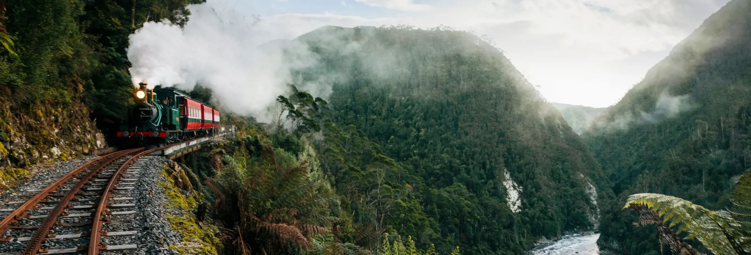 A vintage steam train rounding a curve on a narrow mountain railway, with white steam billowing into a lush gorge of ferns and moss-covered cliffs, a rushing river below and soft light filtering through the mist ahead.