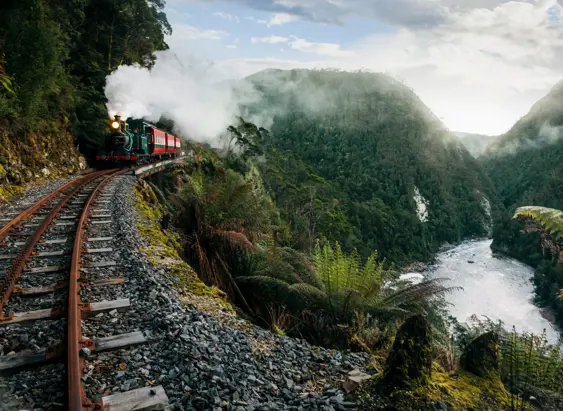 A vintage steam train rounding a curve on a narrow mountain railway, with white steam billowing into a lush gorge of ferns and moss-covered cliffs, a rushing river below and soft light filtering through the mist ahead.